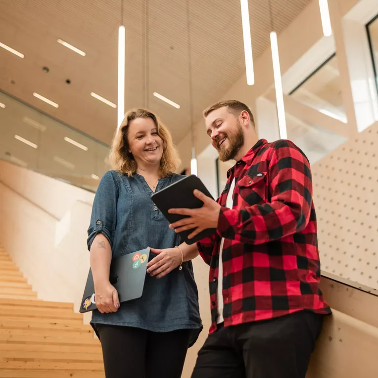 Two smiling colleagues on a wooden staircase looking at a tablet together